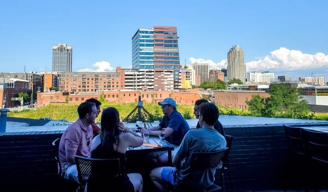 Um grupo de pessoas sentadas em uma mesa no Wye Hill Kitchen & Brewing com o horizonte de Raleigh, Carolina do Norte, ao fundo